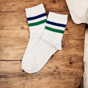May include: A pair of white crew socks with a blue and green stripe design near the top. The socks are made of a soft material and are displayed on a wooden surface.