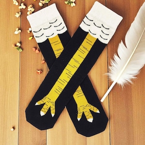 Könnte beinhalten: Ein Paar schwarze kniehohe Socken mit einem gelben Hühnerbein-Design. Die Socken haben weiße Oberteile mit einem gewellten Muster und gelbe Beine mit detaillierten Linien. Auf einer Holzoberfläche mit dekorativen Elementen.