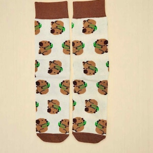 Op de afbeelding: Een paar witte sokken met een bruine boord en teen. De sokken zijn versierd met een herhalend patroon van bruine capibara's met groene slakragen. Een leuk en eigenzinnig ontwerp.