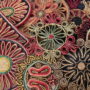 May include: A collection of circular and geometric coasters crafted from coiled, woven fibres in vibrant shades of red, navy blue, olive green, and golden yellow. Each piece features intricate, symmetrical floral and scrollwork patterns.