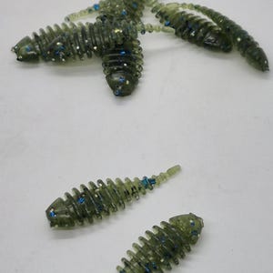 May include: Several green soft plastic fishing lures with a segmented body design and blue glitter accents are scattered on a white surface. The lures are designed to mimic the appearance of small aquatic creatures.
