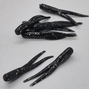 May include: Several black fishing lures with gold flecks are scattered across a white surface. Each lure has a segmented body and multiple tentacle-like appendages. The lures are designed to attract fish.