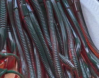 May include: A close-up shot of a pile of soft plastic fishing lures. The lures are a translucent red and green color, with a ribbed body design. The lures are designed to mimic the appearance of worms.