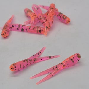May include: A collection of soft plastic fishing lures in a vibrant pink and orange color scheme. The lures have a segmented body design with multiple appendages and dark brown speckles. They are designed to mimic small aquatic creatures.