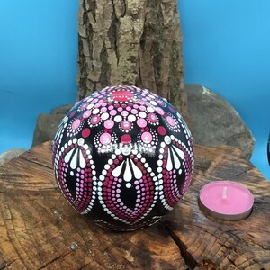 Op de afbeelding: Een zwarte bol met een roze en wit mandala erop geschilderd, rustend op een boomstronk met een roze theelichtje ernaast.
