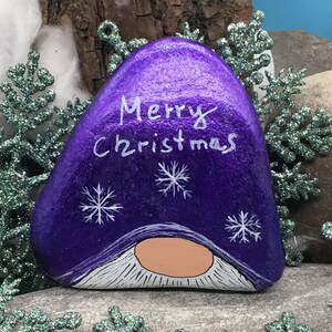 May include: Purple painted rock with a white gnome face and the words "Merry Christmas" in white paint.