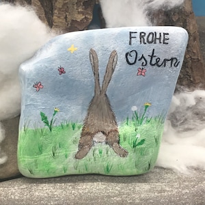 May include: A painted rock with a blue background, a brown bunny rabbit with a white tail, and the text "FROHE OSTERN" in black. The rock is surrounded by green grass and yellow flowers.