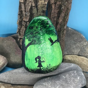 K&ouml;nnte beinhalten: Handbemalter Stein mit der Darstellung eines M&auml;dchens in einem Wald mit einem leuchtenden Glas, einem Baum und einem Vogel. Der Stein ist gr&uuml;n mit wei&szlig;en Akzenten und schwarzen Umrissen.