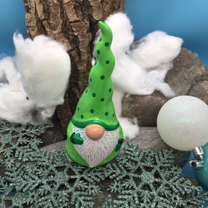 May include: A green gnome with white beard and white polka dots on its hat. The gnome is sitting on a bed of green glitter snowflakes.