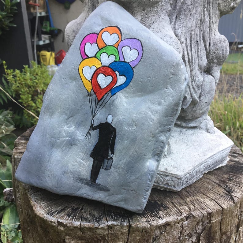 May include: Hand-painted stone art featuring a figure in a black coat holding a bunch of colorful balloons with heart-shaped centers. The balloons are in shades of red, yellow, orange, green, blue, and purple. The stone is gray and has a textured surface.