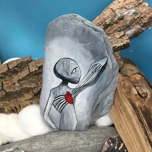 K&ouml;nnte beinhalten: Ein handbemalter grauer Stein zeigt eine au&szlig;erirdische Figur mit einem roten Herzen und freiliegendem Brustkorb. Die Figur streckt einen Arm aus. Das Kunstwerk befindet sich vor einem blauen Hintergrund mit Treibholz und anderen Steinen.