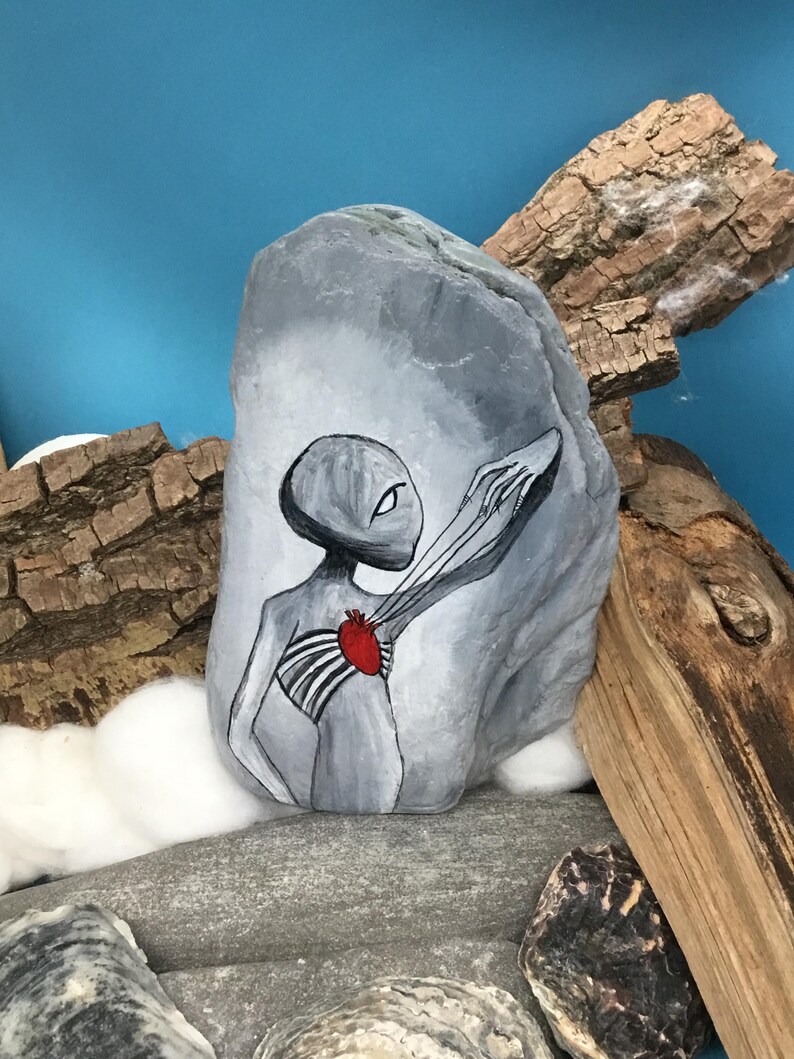 K&ouml;nnte beinhalten: Ein handbemalter grauer Stein zeigt eine au&szlig;erirdische Figur mit einem roten Herzen und freiliegendem Brustkorb. Die Figur streckt einen Arm aus. Das Kunstwerk befindet sich vor einem blauen Hintergrund mit Treibholz und anderen Steinen.