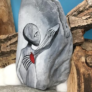 K&ouml;nnte beinhalten: Ein handbemalter grauer Stein zeigt eine Alienfigur mit einem roten Herzen und Rippendetails. Der Arm des Aliens erstreckt sich nach au&szlig;en. Das Kunstwerk ist vor einem blauen Hintergrund mit Holzakzenten platziert und pr&auml;sentiert ein einzigartiges dekoratives Kunstwerk.