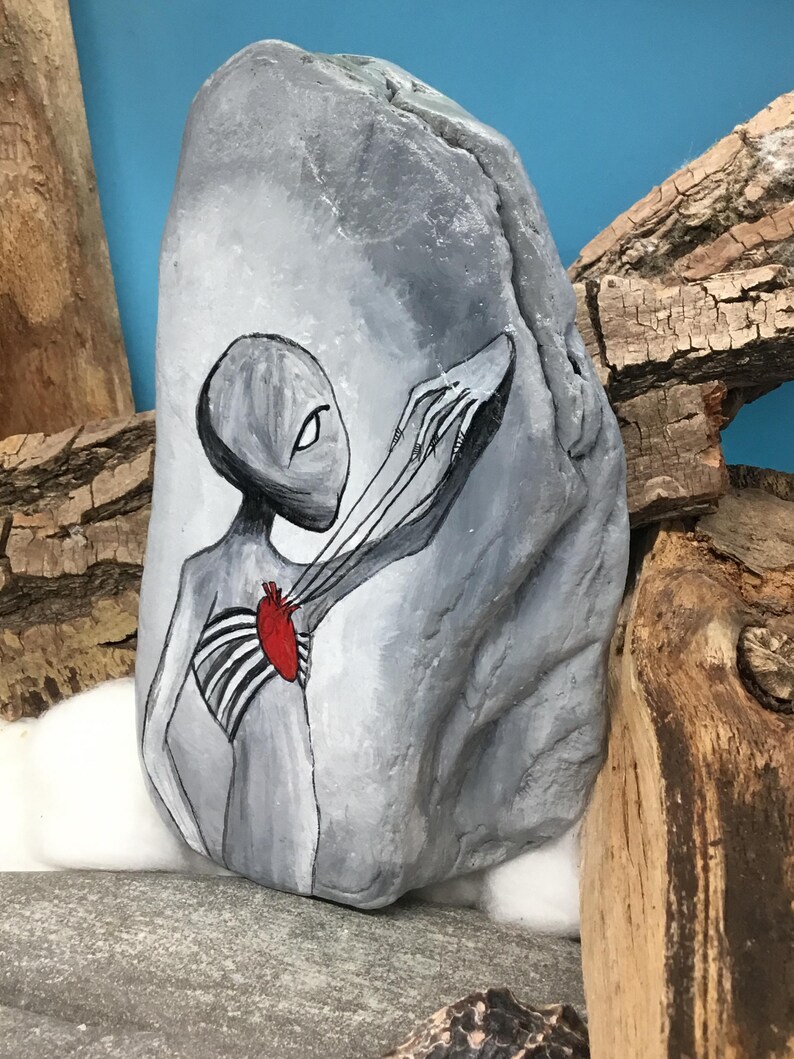 K&ouml;nnte beinhalten: Ein handbemalter grauer Stein zeigt eine Alienfigur mit einem roten Herzen und Rippendetails. Der Arm des Aliens erstreckt sich nach au&szlig;en. Das Kunstwerk ist vor einem blauen Hintergrund mit Holzakzenten platziert und pr&auml;sentiert ein einzigartiges dekoratives Kunstwerk.