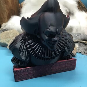 May include: A black 3D-printed bust of a sinister clown with a ruffled collar, resting on a burgundy base. The clown has a bald head and a menacing grin. The background features a blue surface and a cloudy backdrop.