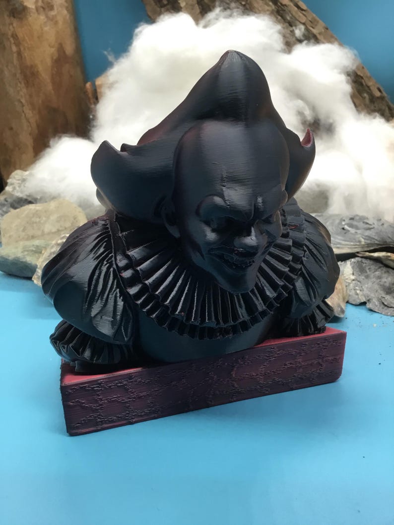 May include: A black 3D-printed bust of a sinister clown with a ruffled collar, resting on a burgundy base. The clown has a bald head and a menacing grin. The background features a blue surface and a cloudy backdrop.