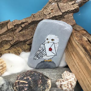May include: A hand-painted, grey stone features a white owl holding a letter with a red wax seal. The owl is perched on a grey rock, set against a blue background with driftwood and seashells. The artwork is detailed and whimsical.