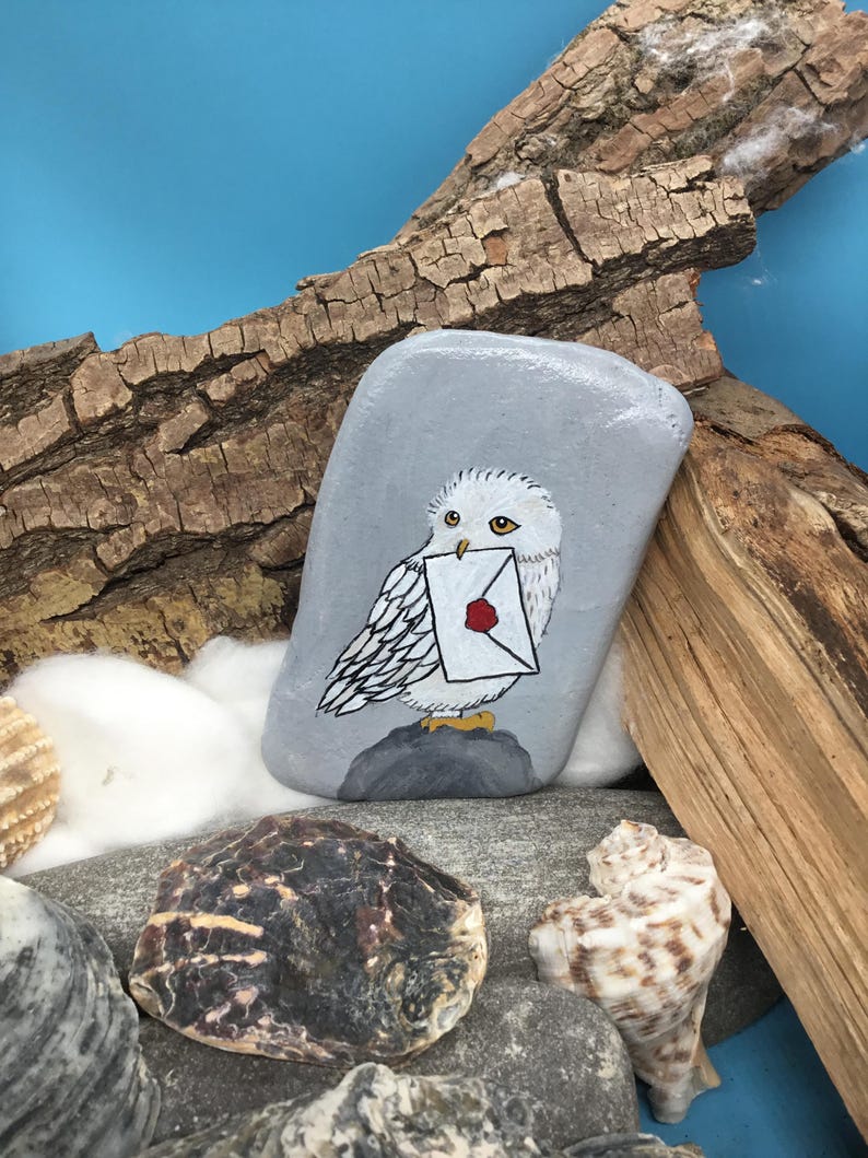 May include: A hand-painted, grey stone features a white owl holding a letter with a red wax seal. The owl is perched on a grey rock, set against a blue background with driftwood and seashells. The artwork is detailed and whimsical.