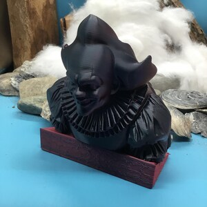 May include: A black bust of a scary clown with a ruffled collar, resting on a rectangular, maroon base. The clown has a sinister grin and a large, ruffled collar. The bust is set against a blue background with rocks, wood, and cotton.