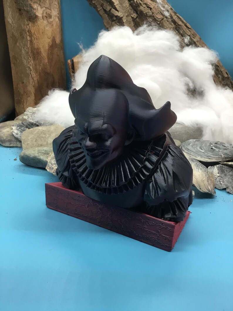 May include: A black bust of a scary clown with a ruffled collar, resting on a rectangular, maroon base. The clown has a sinister grin and a large, ruffled collar. The bust is set against a blue background with rocks, wood, and cotton.