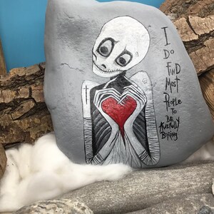 May include: A hand-painted gray rock featuring a skeleton figure holding a red heart. The skeleton has large black eyes and is painted in black and white. Text on the rock reads, "I do find most people to be awfully boring."