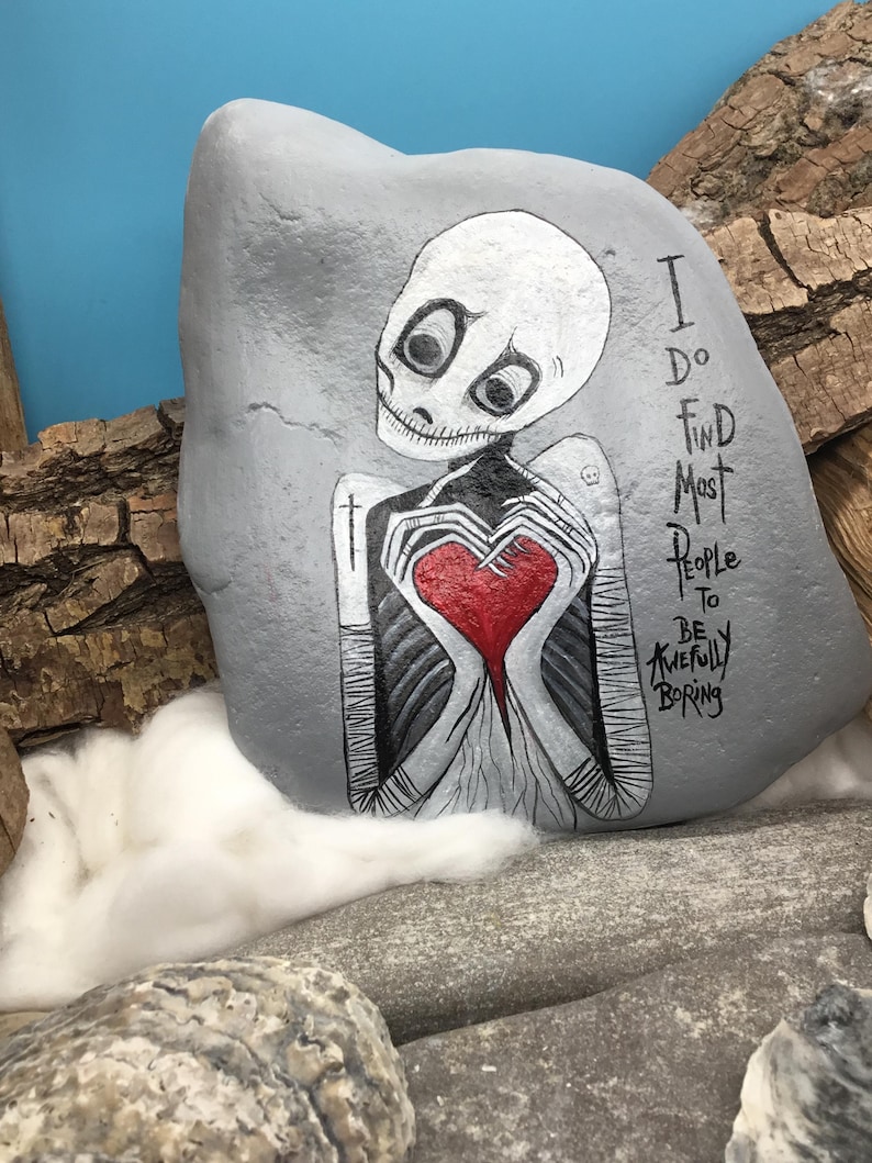 May include: A hand-painted gray rock featuring a skeleton figure holding a red heart. The skeleton has large black eyes and is painted in black and white. Text on the rock reads, "I do find most people to be awfully boring."