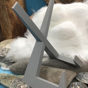 May include: A grey metal display stand with an X-shaped design. The stand is made of a textured material and is designed to hold a small object. The stand is resting on a bed of rocks and cotton.
