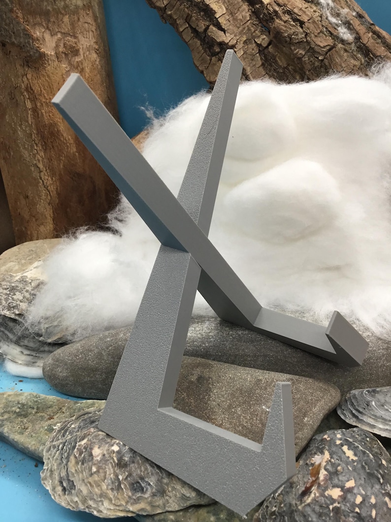 May include: A grey metal display stand with an X-shaped design. The stand is made of a textured material and is designed to hold a small object. The stand is resting on a bed of rocks and cotton.