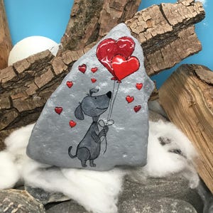 May include: A hand-painted rock with a gray background featuring a cartoon dog holding two red heart-shaped balloons.  The dog is surrounded by smaller red hearts.