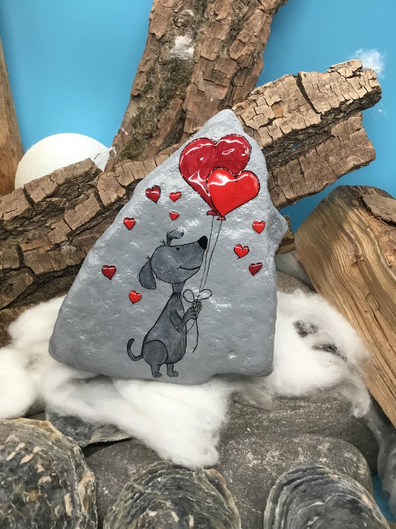 May include: A hand-painted rock with a gray background featuring a cartoon dog holding two red heart-shaped balloons.  The dog is surrounded by smaller red hearts.