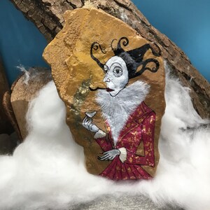 Op de afbeelding: Een handbeschilderde steen toont een grillige figuur in een rood-gouden jas met een witte kraag met ruches, die een klein kopje vasthoudt. De figuur heeft een bleek gezicht, zwart haar en staat tegen een goudbruine achtergrond.