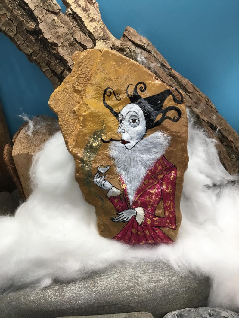 Op de afbeelding: Een handbeschilderde steen toont een grillige figuur in een rood-gouden jas met een witte kraag met ruches, die een klein kopje vasthoudt. De figuur heeft een bleek gezicht, zwart haar en staat tegen een goudbruine achtergrond.