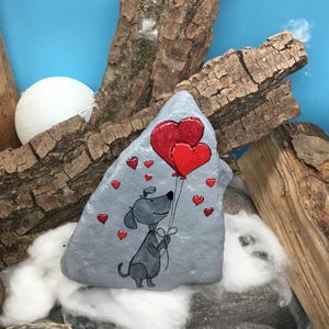 May include: A gray painted rock with a cartoon dog holding two red heart-shaped balloons. The dog is smiling and has a black nose and eyes. There are smaller red hearts scattered around the dog and balloons.
