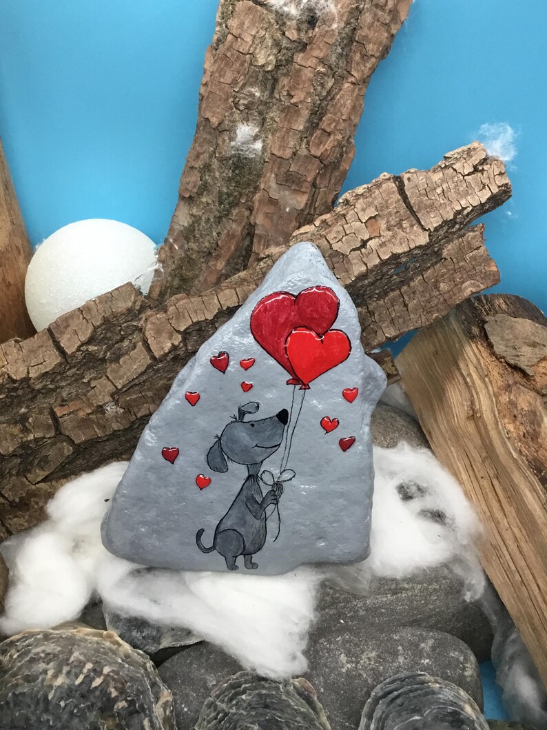 May include: A gray painted rock with a cartoon dog holding two red heart-shaped balloons. The dog is smiling and has a black nose and eyes. There are smaller red hearts scattered around the dog and balloons.
