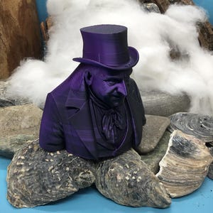 May include: A purple bust of a man in a top hat and suit, set on rocks and oyster shells. The background features cotton and driftwood. The figure has a serious expression. The bust is a decorative item.