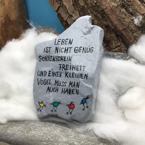 May include: A light gray painted stone featuring black hand-lettered German text that reads LEBEN IST NICHT GENUG SONNENSCHEIN FREIHEIT UND EINEN KLEINEN VOGEL MUSS MAN AUCH HABEN, accompanied by four small, colorful bird illustrations in yellow, green, red, and blue.