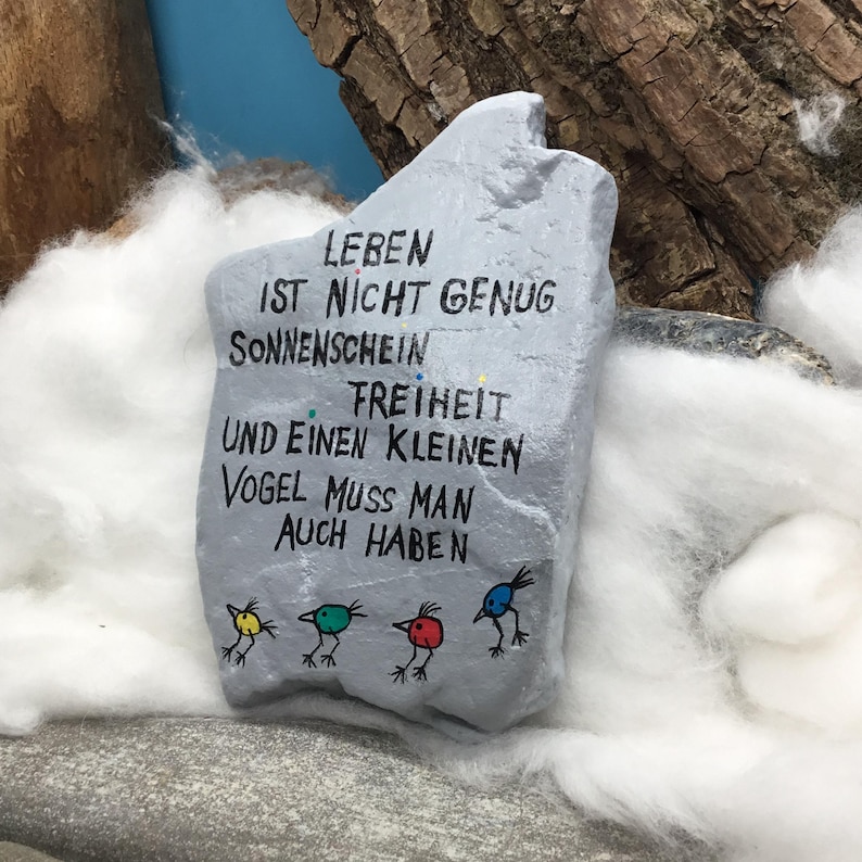 May include: A light gray painted stone featuring black hand-lettered German text that reads LEBEN IST NICHT GENUG SONNENSCHEIN FREIHEIT UND EINEN KLEINEN VOGEL MUSS MAN AUCH HABEN, accompanied by four small, colorful bird illustrations in yellow, green, red, and blue.