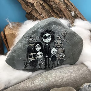 Puede incluir: Una piedra gris lisa que presenta una ilustraci&oacute;n detallada pintada a mano de un grupo de personajes estilizados y sombr&iacute;os con ojos grandes y rasgos oscuros. La obra utiliza pintura blanca y negra para representar a las diversas figuras.