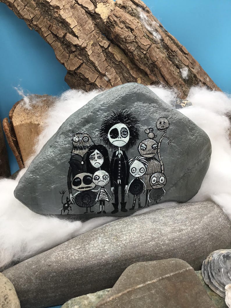 Puede incluir: Una piedra gris lisa que presenta una ilustraci&oacute;n detallada pintada a mano de un grupo de personajes estilizados y sombr&iacute;os con ojos grandes y rasgos oscuros. La obra utiliza pintura blanca y negra para representar a las diversas figuras.