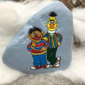 Könnte beinhalten: Ein handbemalter, hellblauer Stein zeigt eine Cartoon-Darstellung von zwei Figuren. Eine Figur ist gelb mit einem gestreiften Hemd und grüner Hose. Die andere Figur ist braun mit einem rot-blau gestreiften Hemd und blauer Hose.