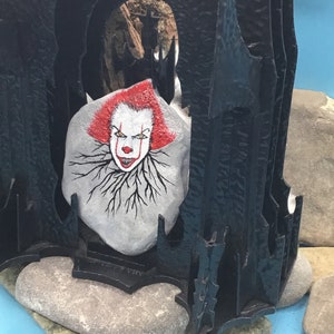 May include: A hand-painted rock with a scary clown face, set inside a black, gothic-style structure. The rock is painted with red hair, white skin, and a black outline. The clown's eyes are yellow, and the mouth is red with sharp teeth. The structure is made of black metal and has a pointed archway.
