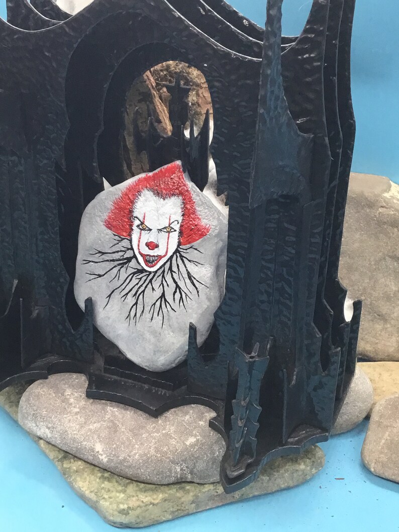 May include: A hand-painted rock with a scary clown face, set inside a black, gothic-style structure. The rock is painted with red hair, white skin, and a black outline. The clown's eyes are yellow, and the mouth is red with sharp teeth. The structure is made of black metal and has a pointed archway.