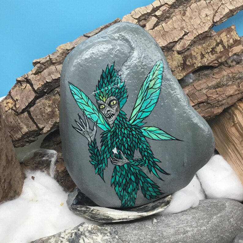 May include: Hand-painted gray rock featuring a green fairy with detailed wings and a menacing expression. The fairy is holding a small white object. The rock is set against a backdrop of wood and blue.
