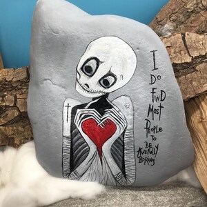 May include: A hand-painted gray rock features a skeletal figure holding a red heart. The figure has large, expressive eyes and is painted in black and white. Text on the rock reads, "I do find most people to be awfully boring."