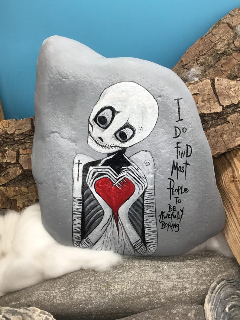 May include: A hand-painted gray rock features a skeletal figure holding a red heart. The figure has large, expressive eyes and is painted in black and white. Text on the rock reads, "I do find most people to be awfully boring."