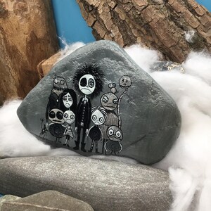 Puede incluir: Una piedra gris lisa con una ilustraci&oacute;n detallada pintada a mano de un grupo de personajes monocrom&aacute;ticos estilizados con ojos grandes y rostros expresivos. La obra muestra una figura central alta rodeada de varias criaturas peque&ntilde;as y variadas.