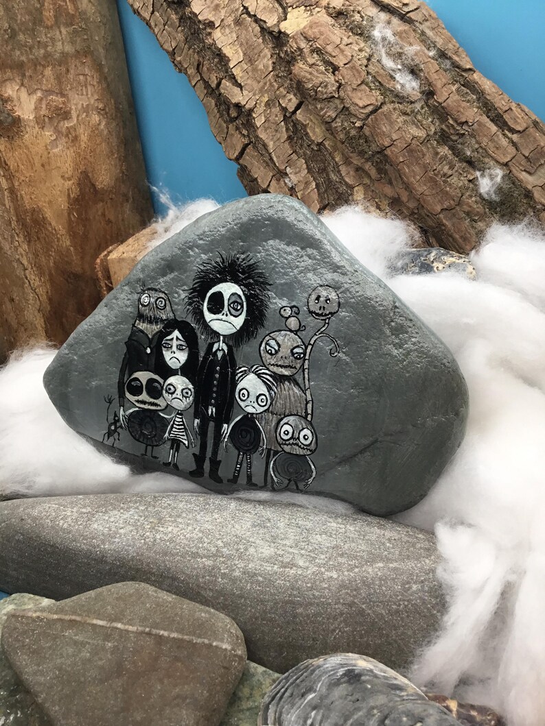 Puede incluir: Una piedra gris lisa con una ilustraci&oacute;n detallada pintada a mano de un grupo de personajes monocrom&aacute;ticos estilizados con ojos grandes y rostros expresivos. La obra muestra una figura central alta rodeada de varias criaturas peque&ntilde;as y variadas.