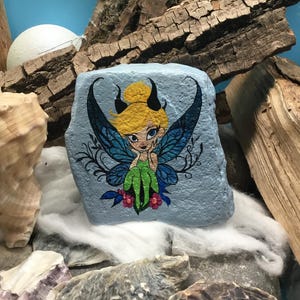 May include: Hand-painted rock art featuring a fairy with blue wings, green dress, and yellow hair. The fairy is sitting on a branch with pink flowers. The rock is painted a light blue color.