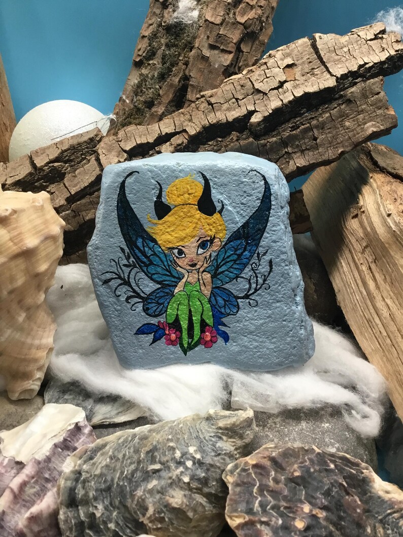 May include: Hand-painted rock art featuring a fairy with blue wings, green dress, and yellow hair. The fairy is sitting on a branch with pink flowers. The rock is painted a light blue color.