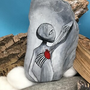 K&ouml;nnte beinhalten: Ein grau bemalter Stein zeigt eine stilisierte Alienfigur mit einem roten Herzen und freiliegendem Brustkorb. Das Kunstwerk ist in Schwarz, Wei&szlig; und Rot gehalten, wobei der Au&szlig;erirdische sich ausstreckt. Der Stein wird auf einer Oberfl&auml;che mit Holz und anderen Steinen ausgestellt.